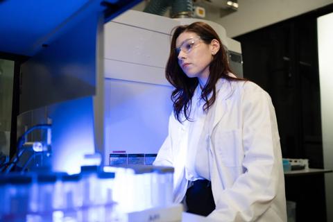 A focused female researcher wearing safety goggles and lab coat operates advanced laboratory equipment illuminated by blue light, ideal for science and technology concepts.