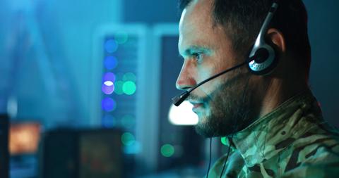 Pan around view of serious commander in camouflage speaking on headset and using computer while working in mission control center during war military & government