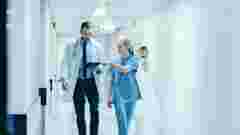 Female Surgeon and Doctor Walk Through Hospital Hallway, They Consult Digital Tablet Computer while Talking about Patient's Health. Modern Bright Hospital with Professional Staff.
