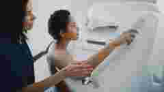 Friendly Female Doctor Explains the Mammogram Procedure to a Topless Latin Female Patient with Curly Hair Undergoing Mammography Scan. Healthy Female Does Cancer Prevention Routine in Hospital Room.