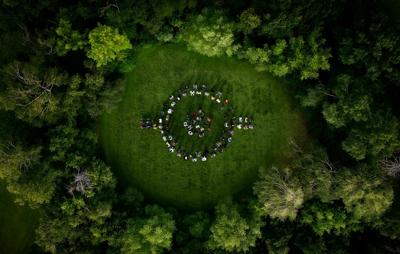 Aerial view of Barco employees forming the Barco logo on a grassy clearing, surrounded by dense green forest. At Barco, we consider sustainability a key part of our DNA.